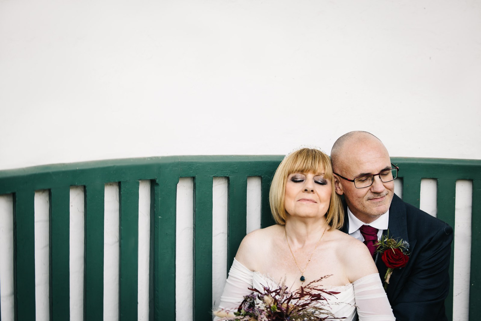 The bride rests her eyes as the couple sit close on a green garden bench