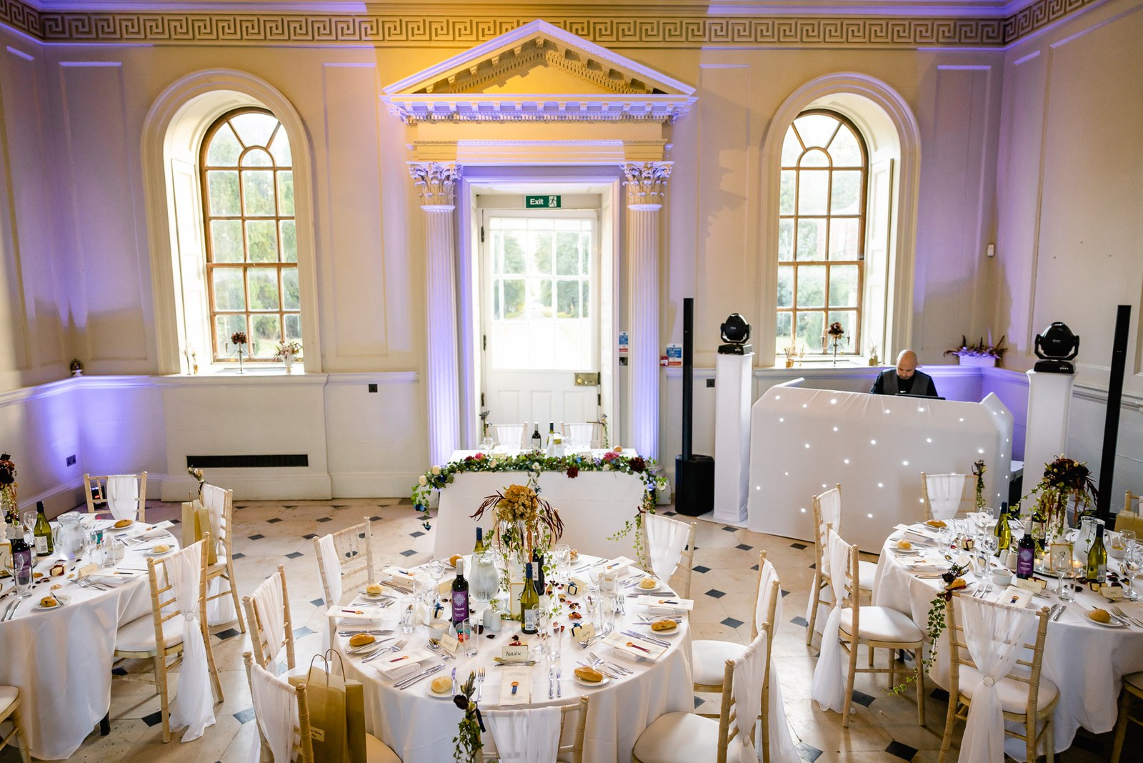 The reception room set with round tables and purple uplighting beneath a gilded cornice