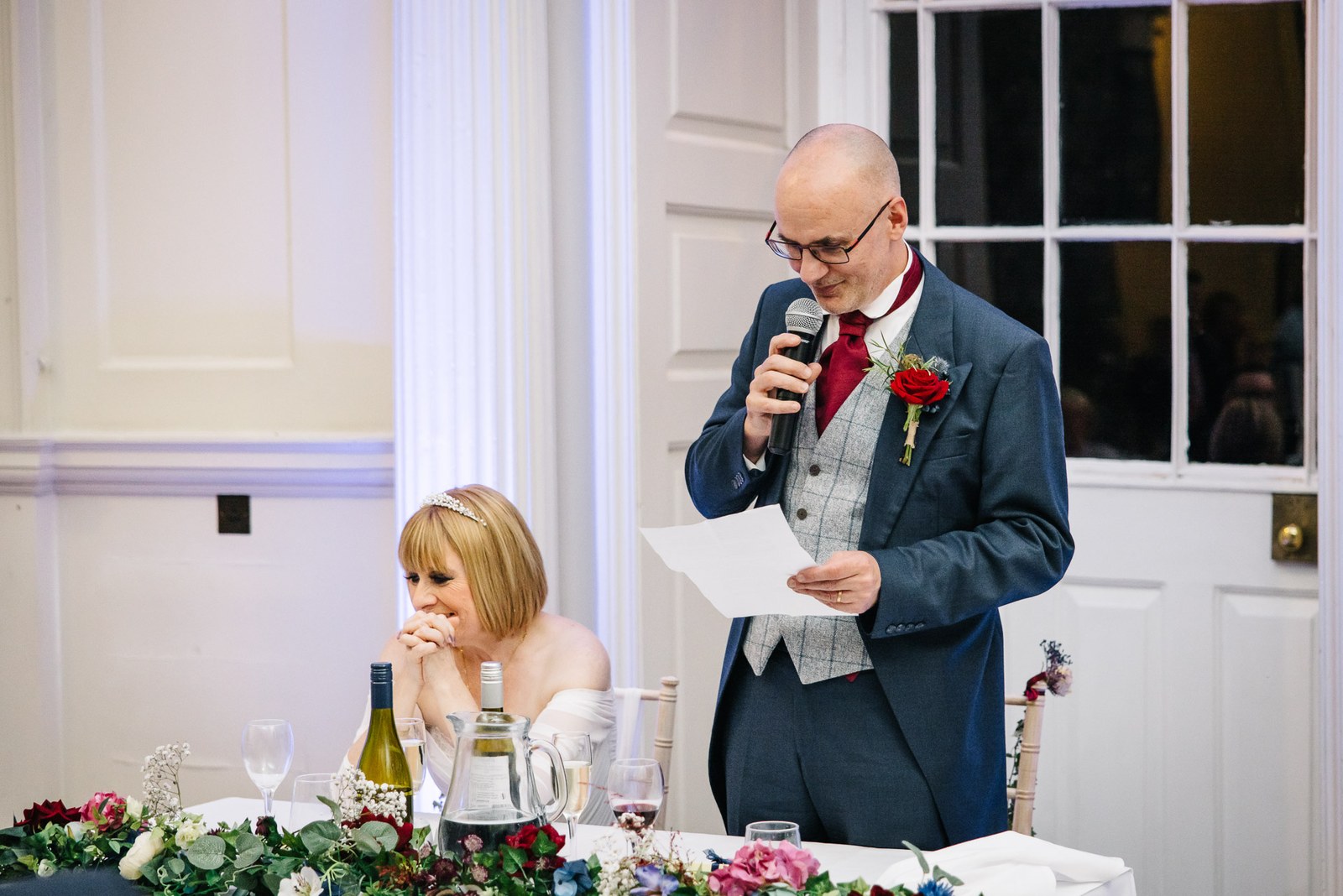 The groom delivers his speech into a microphone as the bride watches from the top table