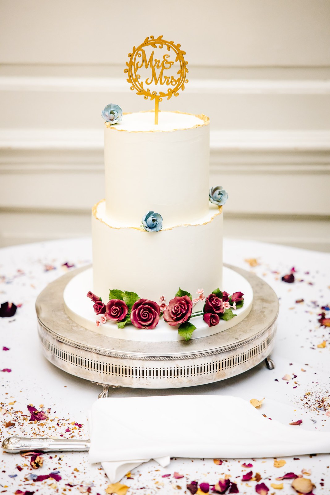 Two-tier white wedding cake with roses and a 'Mr & Mrs' topper amid scattered petals