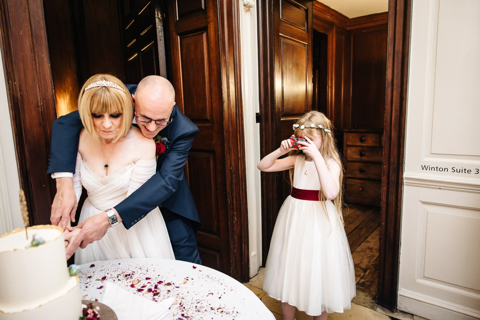 The couple cut their wedding cake as a young guest photographs them from the doorway