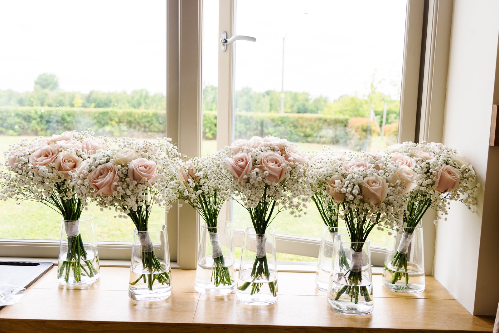 Bridesmaids' bouquets lined up in vases on a windowsill with fields beyond