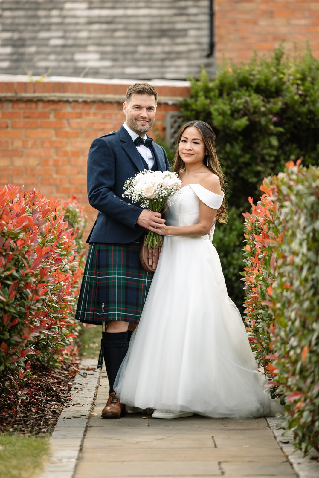 The couple on the paved path between the brick wall and a red-leaved hedge