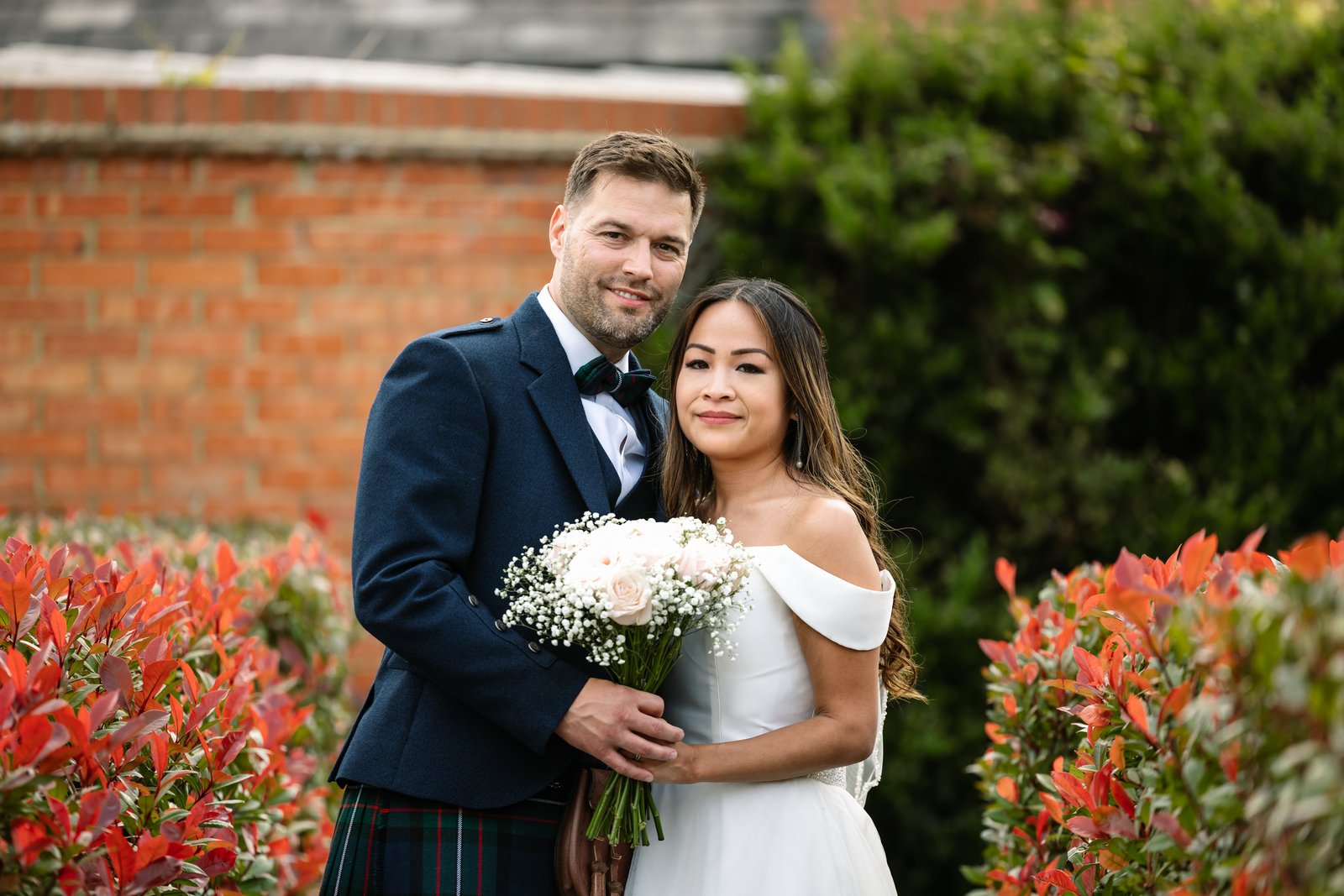 The couple standing together against the brick wall and red hedge
