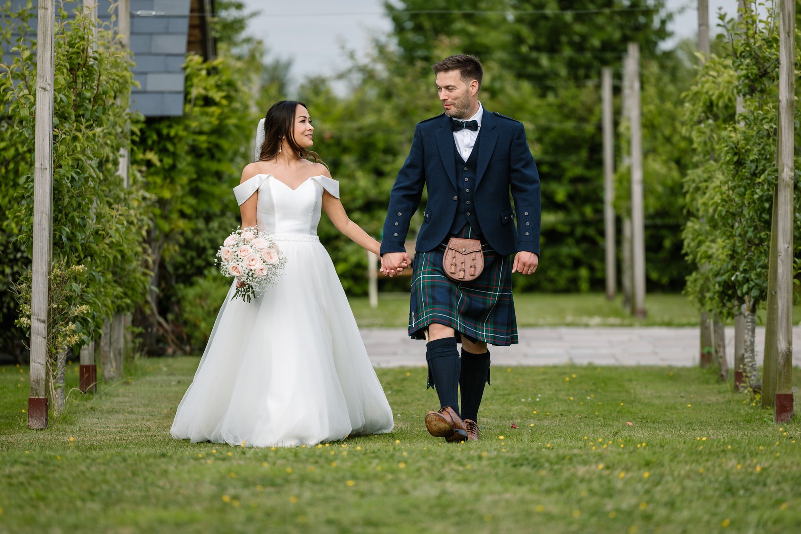 The couple walking hand in hand along a grass path between the rows at Cider Mill Barns