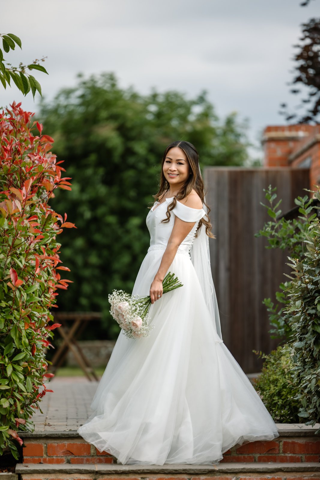 The bride on the brick steps beside a red-leaved hedge at Cider Mill Barns