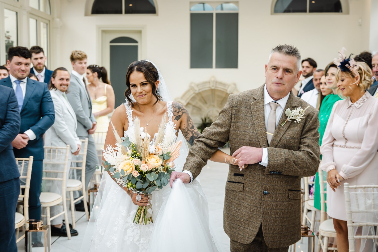 The bride walks down the aisle on her father's arm as guests stand
