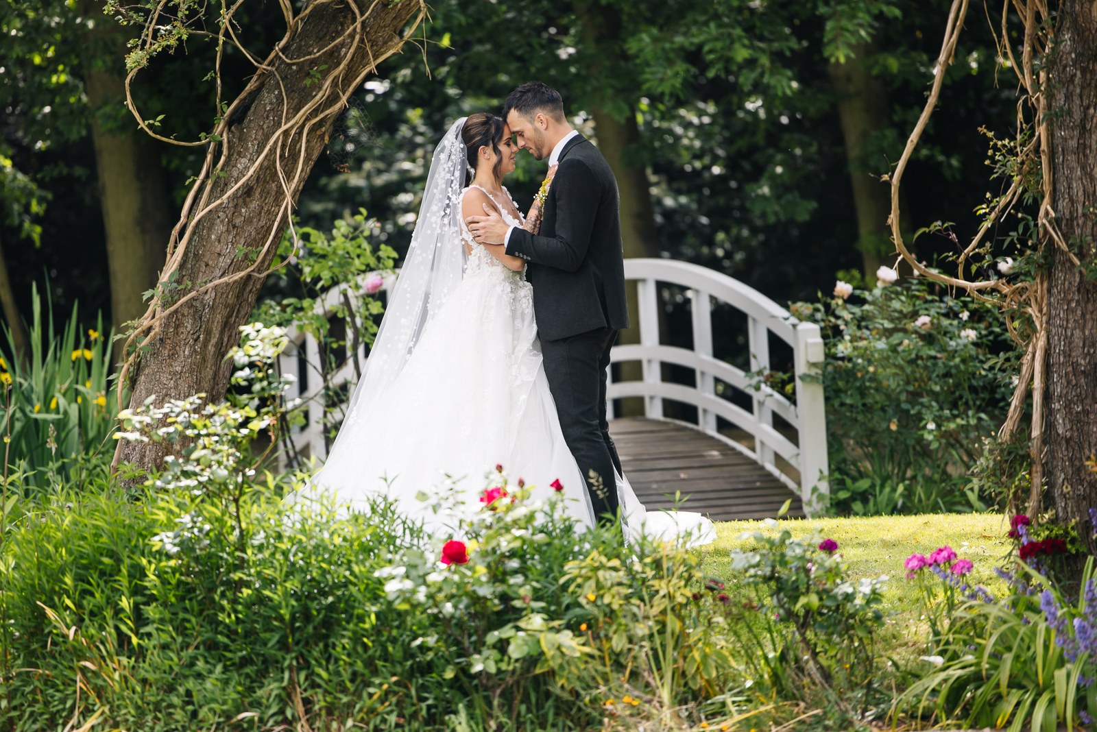 Bride and groom embrace beneath a tree beside the garden bridge