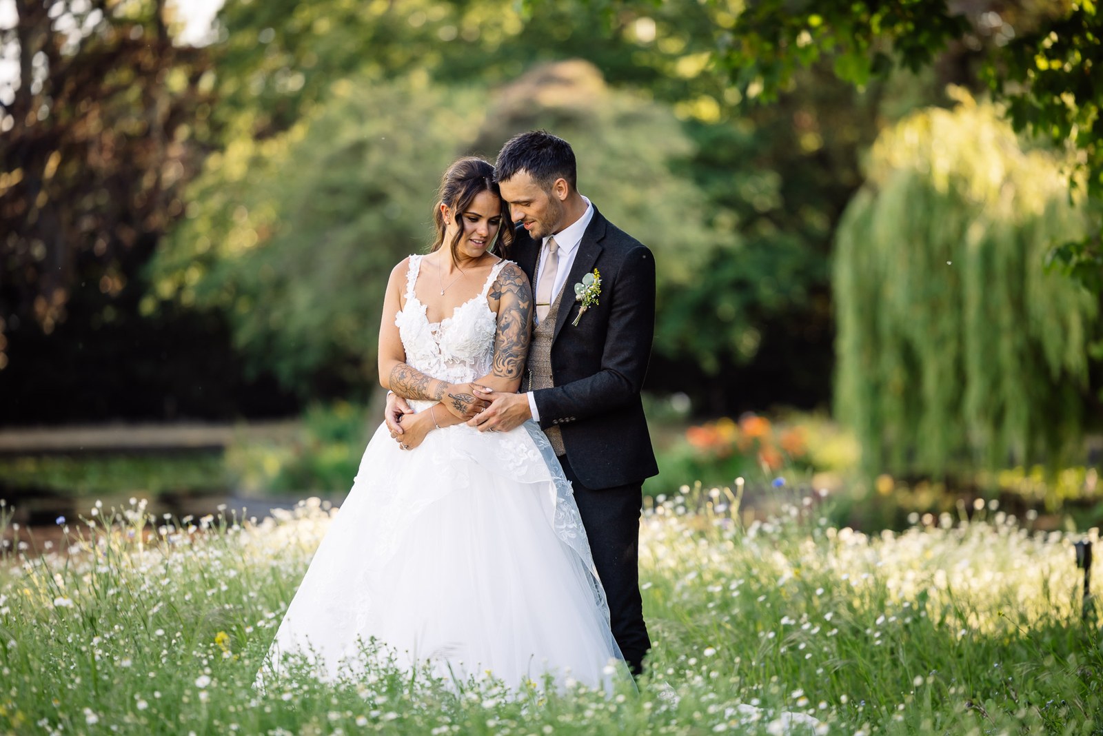 Bride and groom hold each other in a daisy-filled meadow by the willows