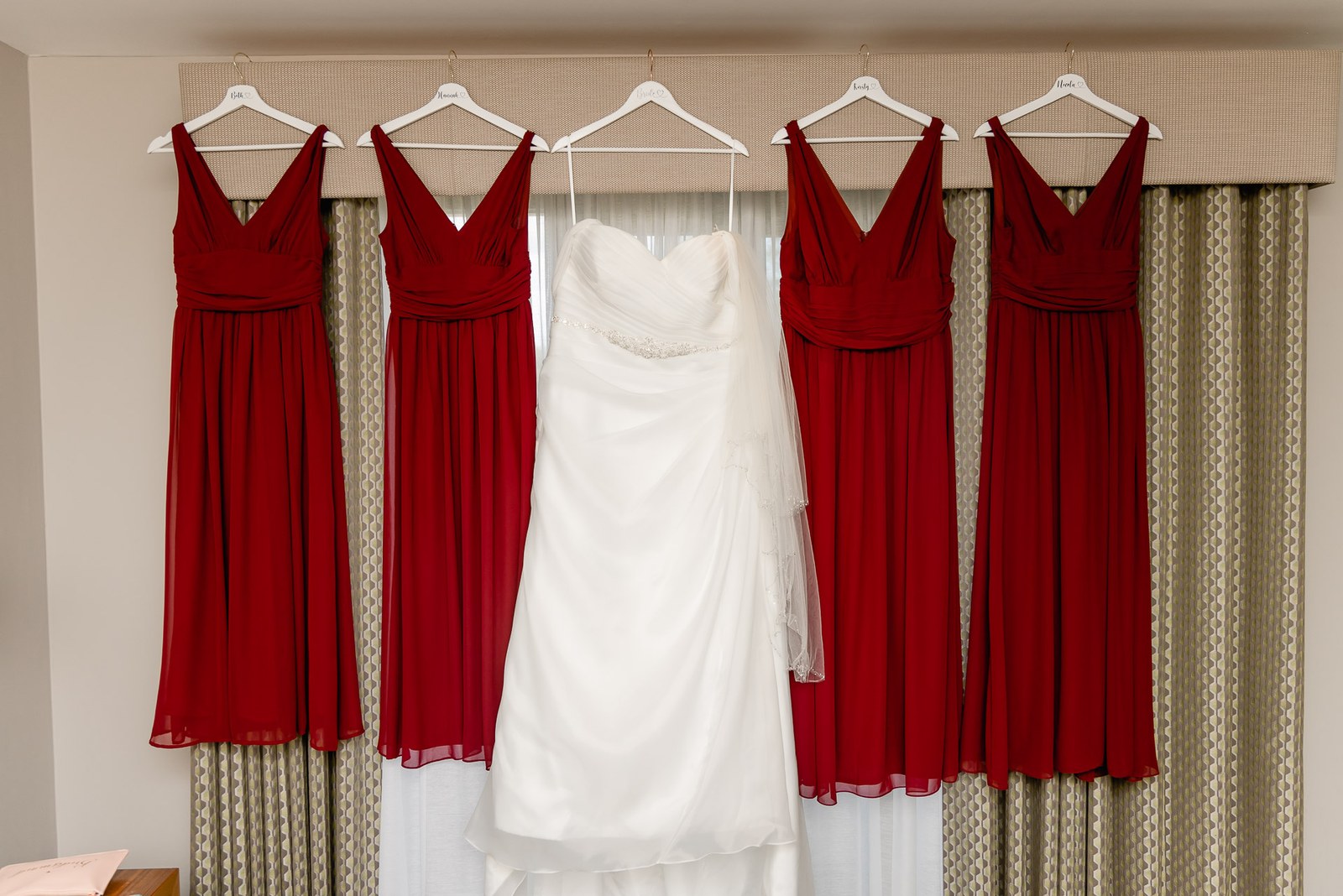 The bride's gown flanked by red bridesmaid dresses hung along a curtain rail