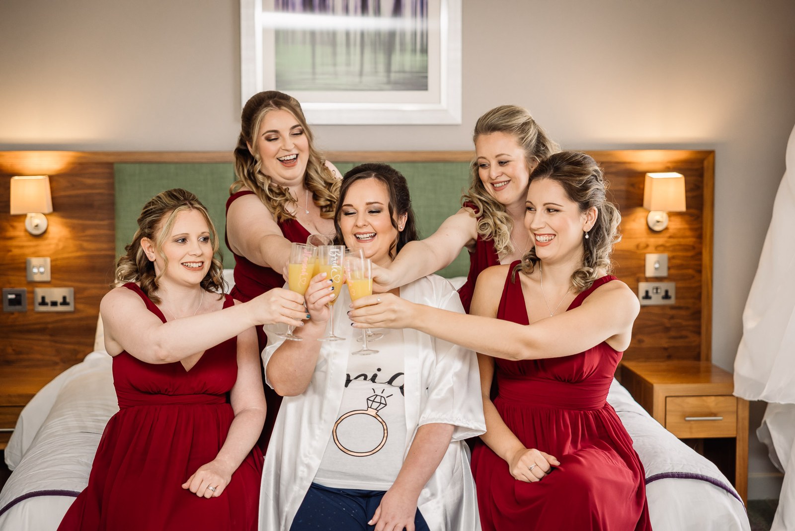 The bride and her bridesmaids in red toast with buck's fizz on the bed