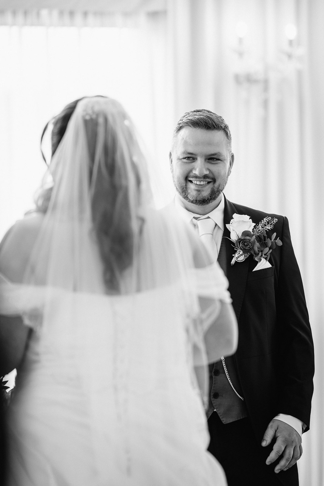 Black-and-white frame of the groom smiling as he faces the veiled bride