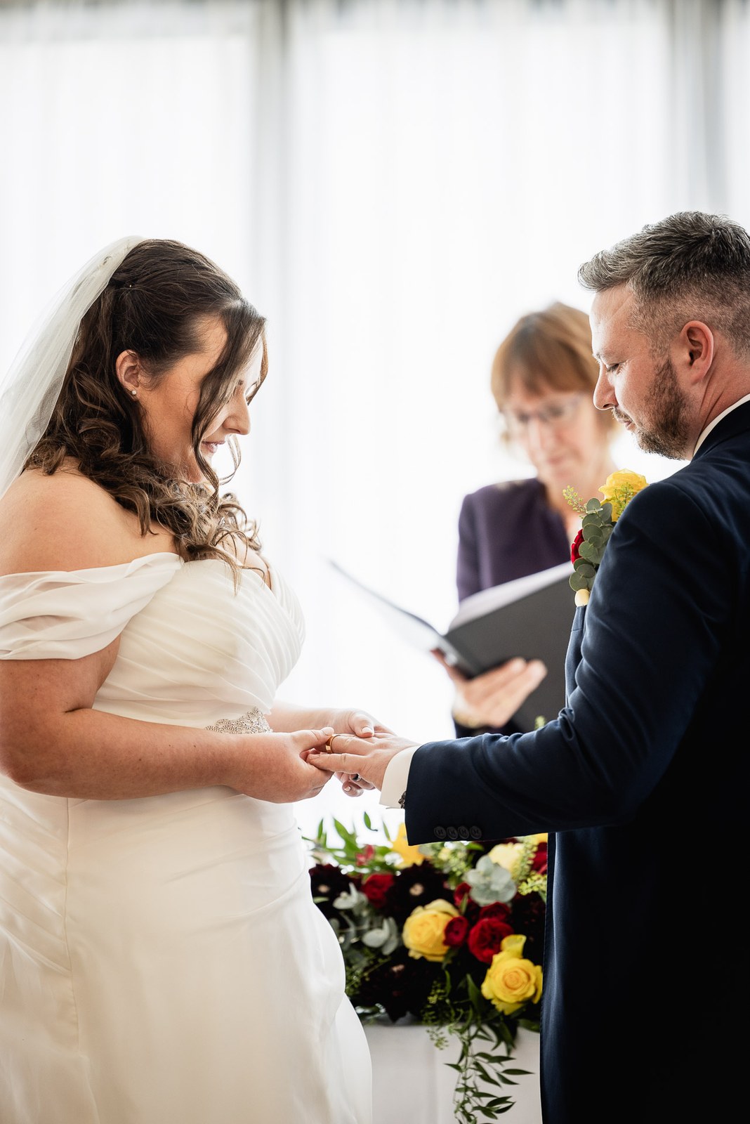 The couple exchange rings before the celebrant, yellow and red roses on the table