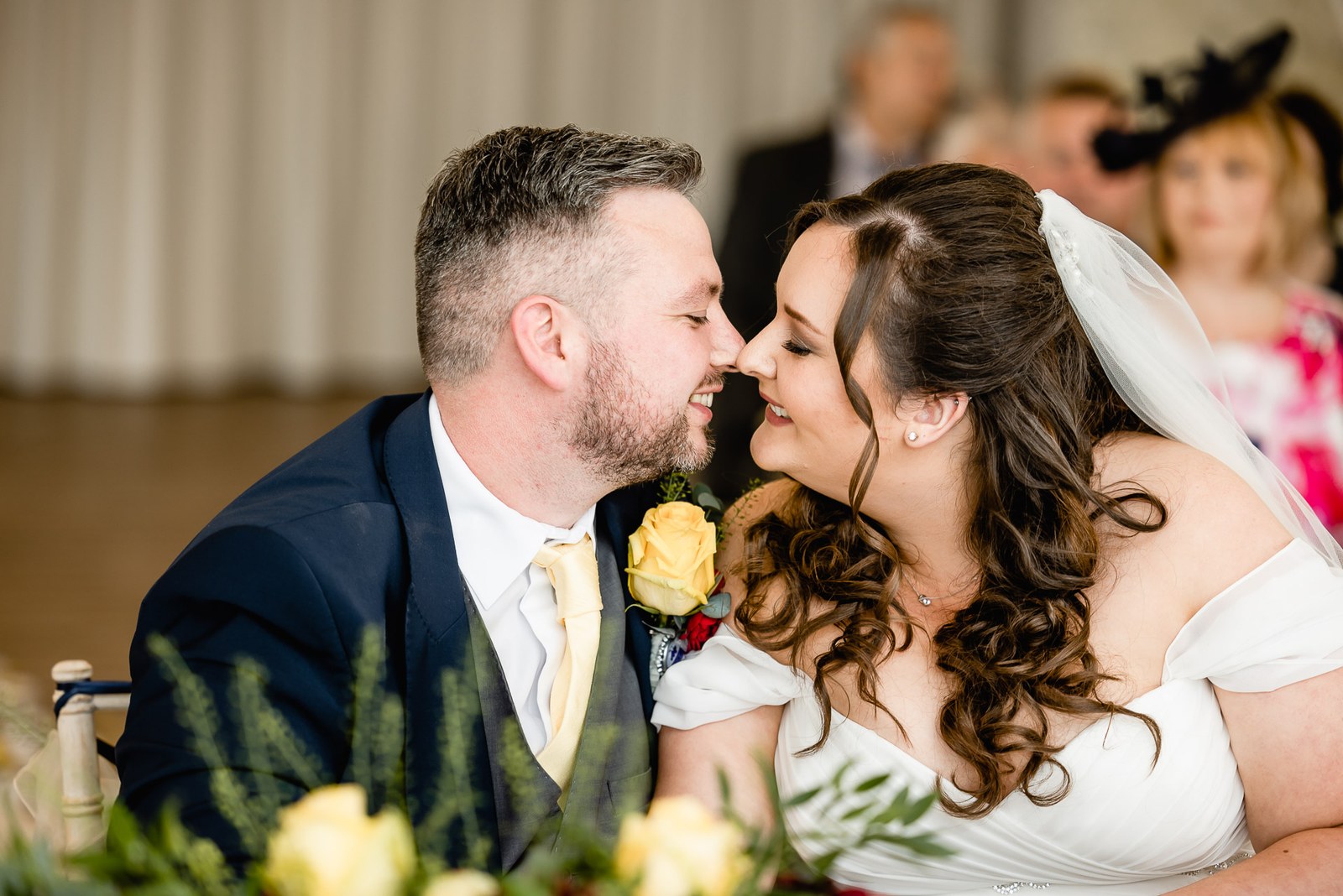 The couple lean their noses together at the table, a yellow rose between them