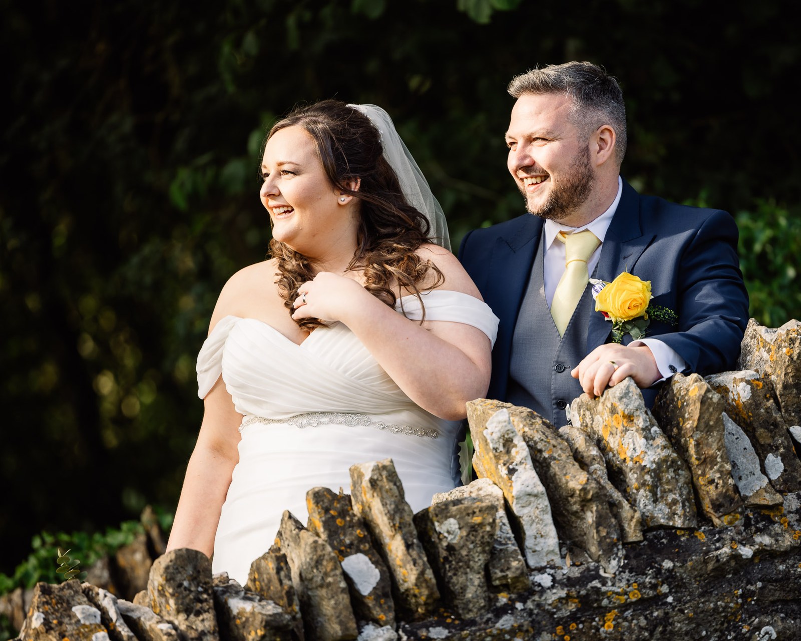 The couple lean on a Cotswold dry-stone wall, both laughing