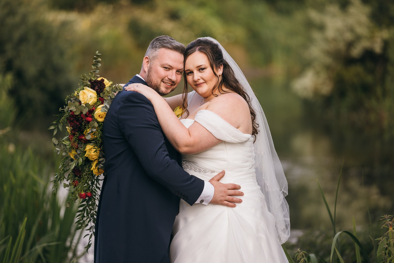 Bride and groom embrace among the lakeside reeds, her bouquet over his shoulder