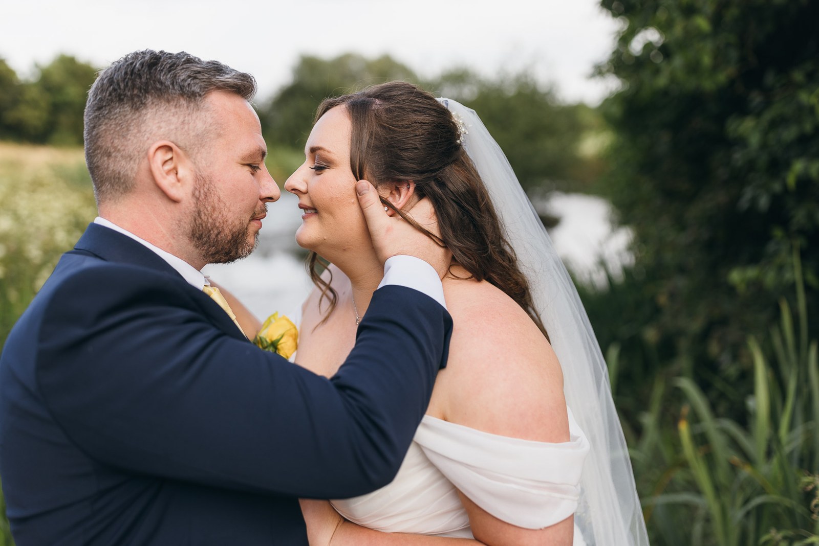 The couple stand nose to nose beside the water in the evening light