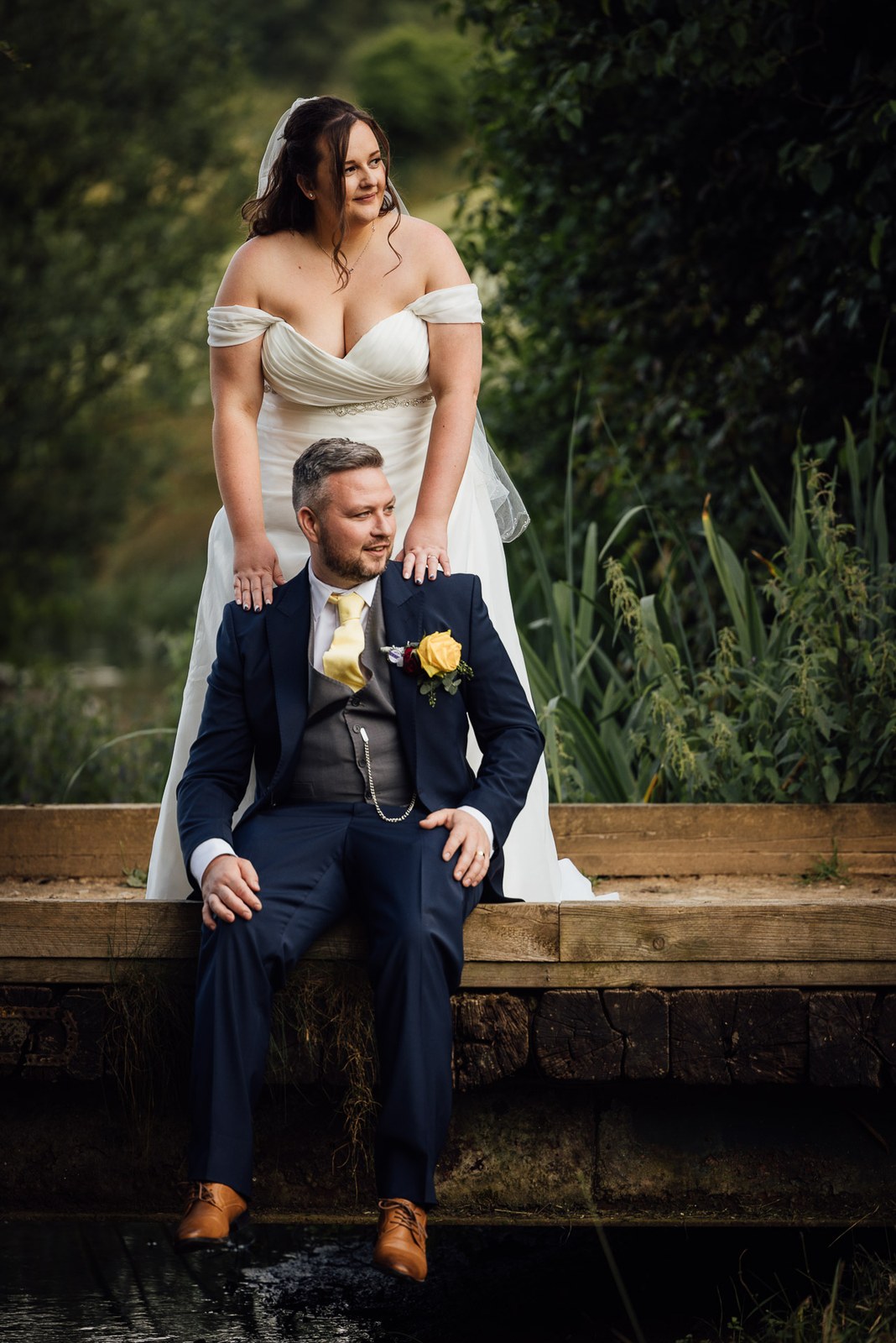 The bride stands behind the groom seated on a wooden jetty over the water