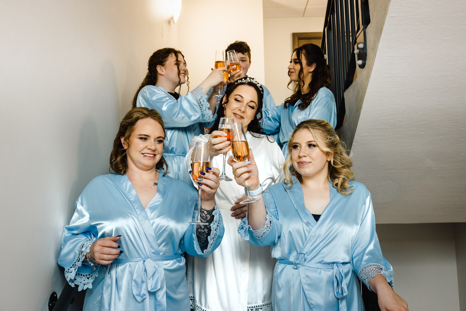 Bridesmaids in pale blue satin robes raising champagne glasses with the bride during morning preparations