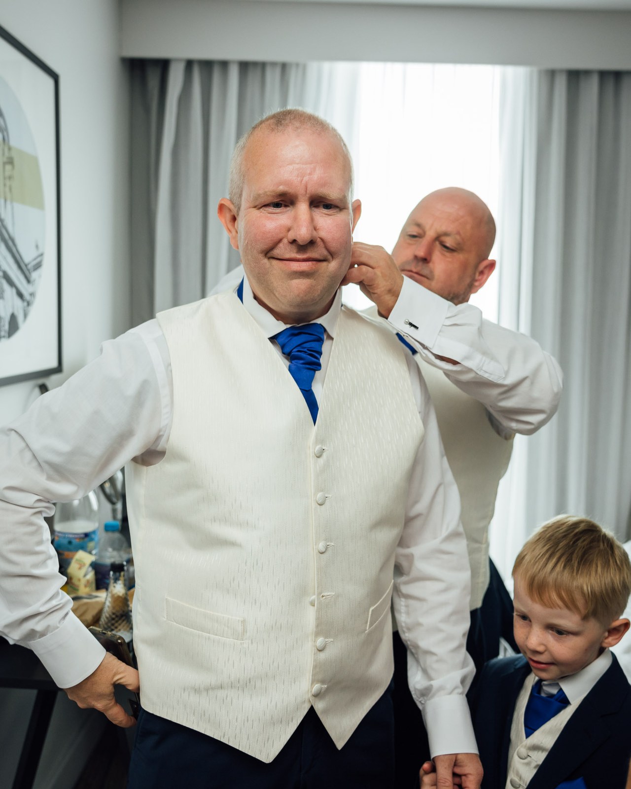 Groom in an ivory waistcoat and blue tie having his collar adjusted, a young page boy beside him