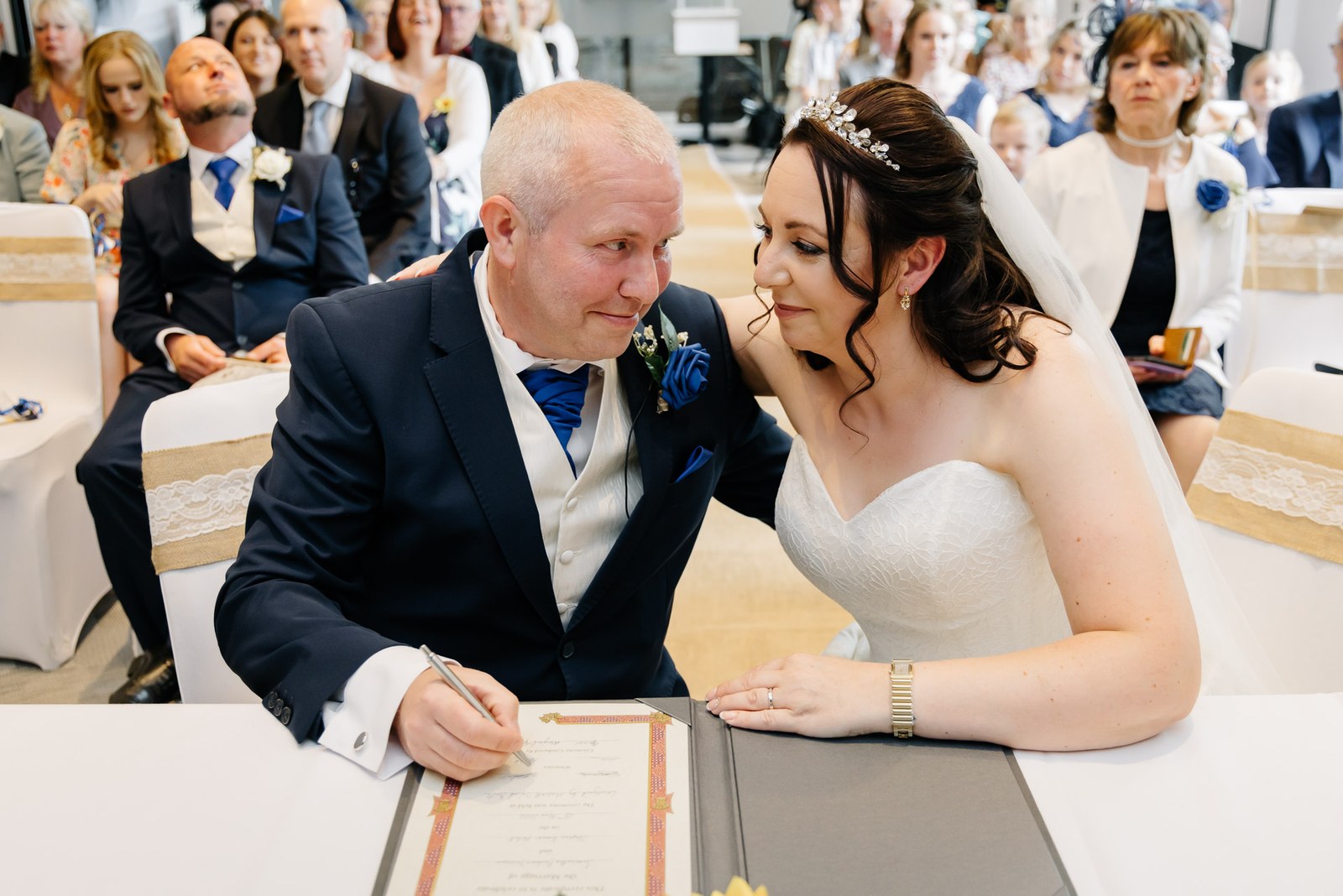Groom signing the marriage register as the bride leans in beside him at the ceremony table