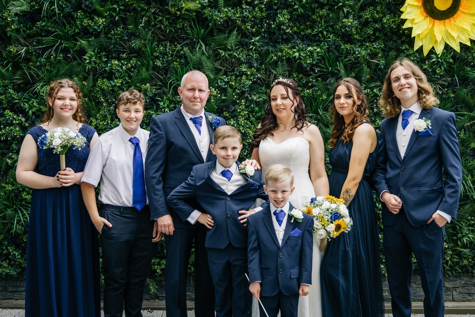 Family group portrait in navy against a green foliage wall, the bride holding her sunflower bouquet