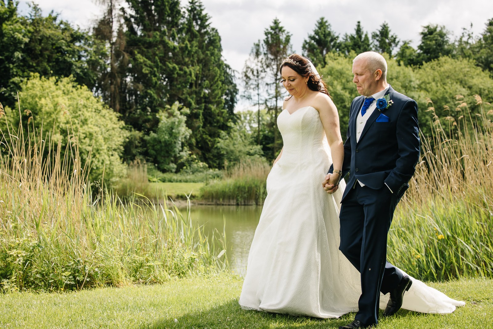 Bride and groom walking on a lawn beside a lake amid summer greenery