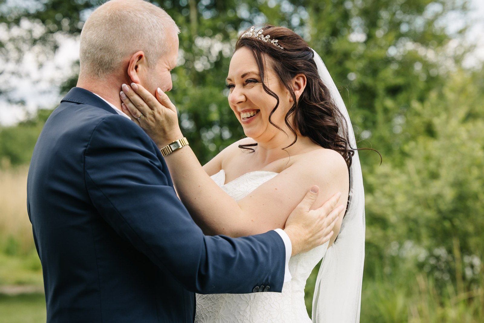 Bride cupping the groom's face as they laugh together among summer greenery