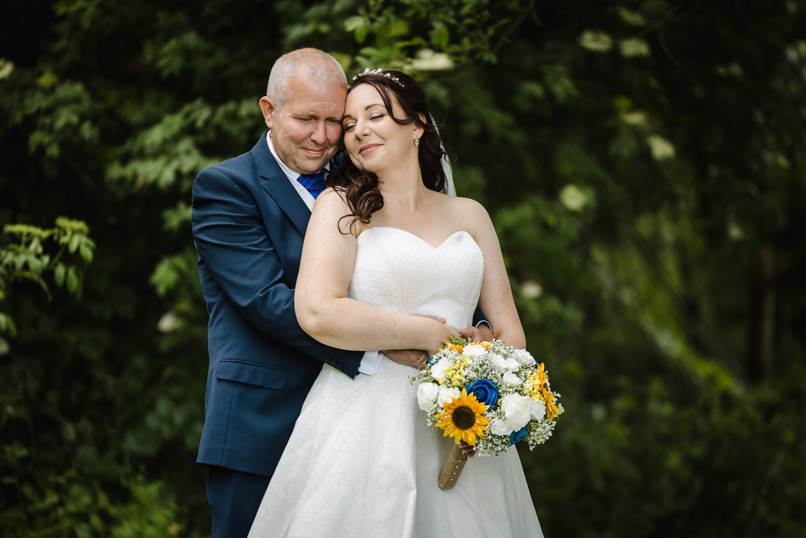 Bride and groom embracing outdoors, she holds a sunflower and blue rose bouquet