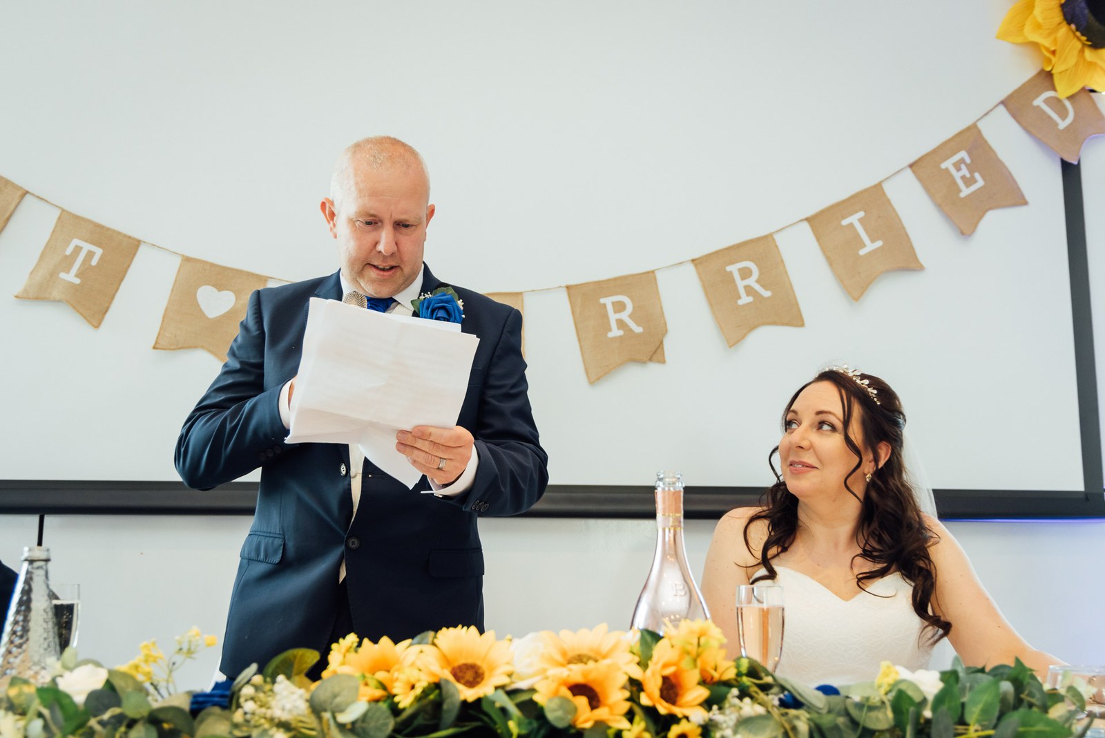 A speech read at the top table beneath 'married' bunting and a sunflower garland