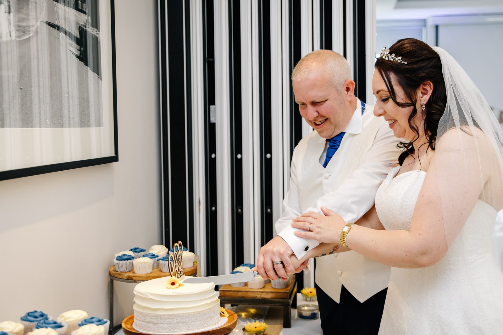 Bride and groom cutting their wedding cake together beside a display of cupcakes