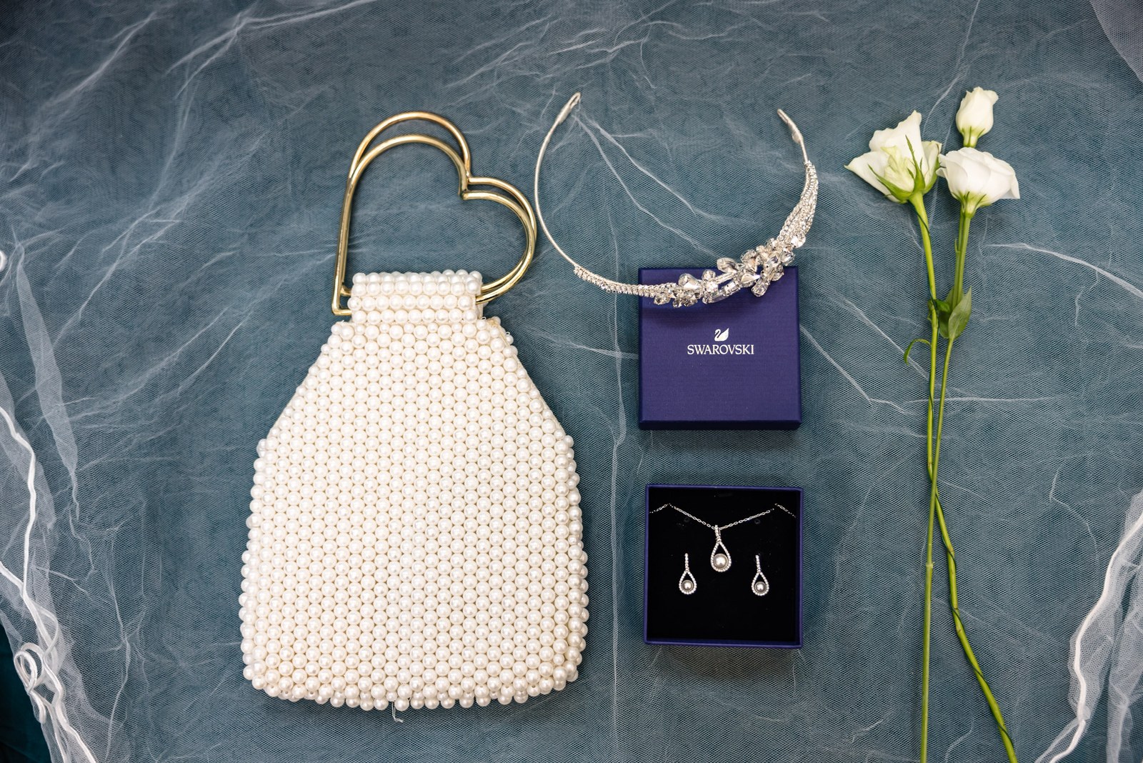 Flat lay of the bride's pearl clutch, tiara and drop earrings beside a white bloom on the veil