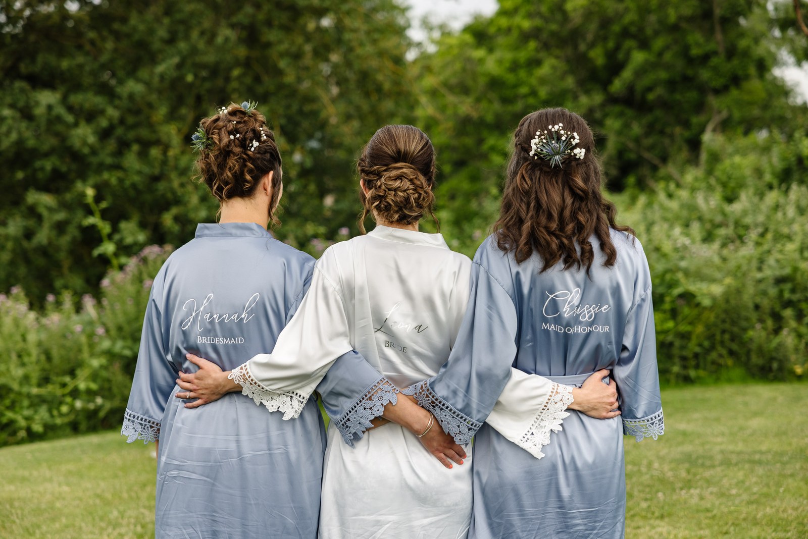 The bride and two bridesmaids in personalised robes seen from behind, arms around each other in the garden