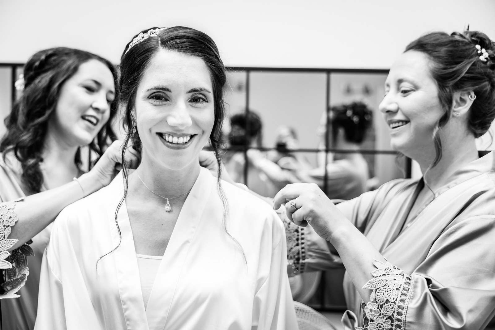 Black-and-white moment of bridesmaids fastening the bride's necklace as she gets ready