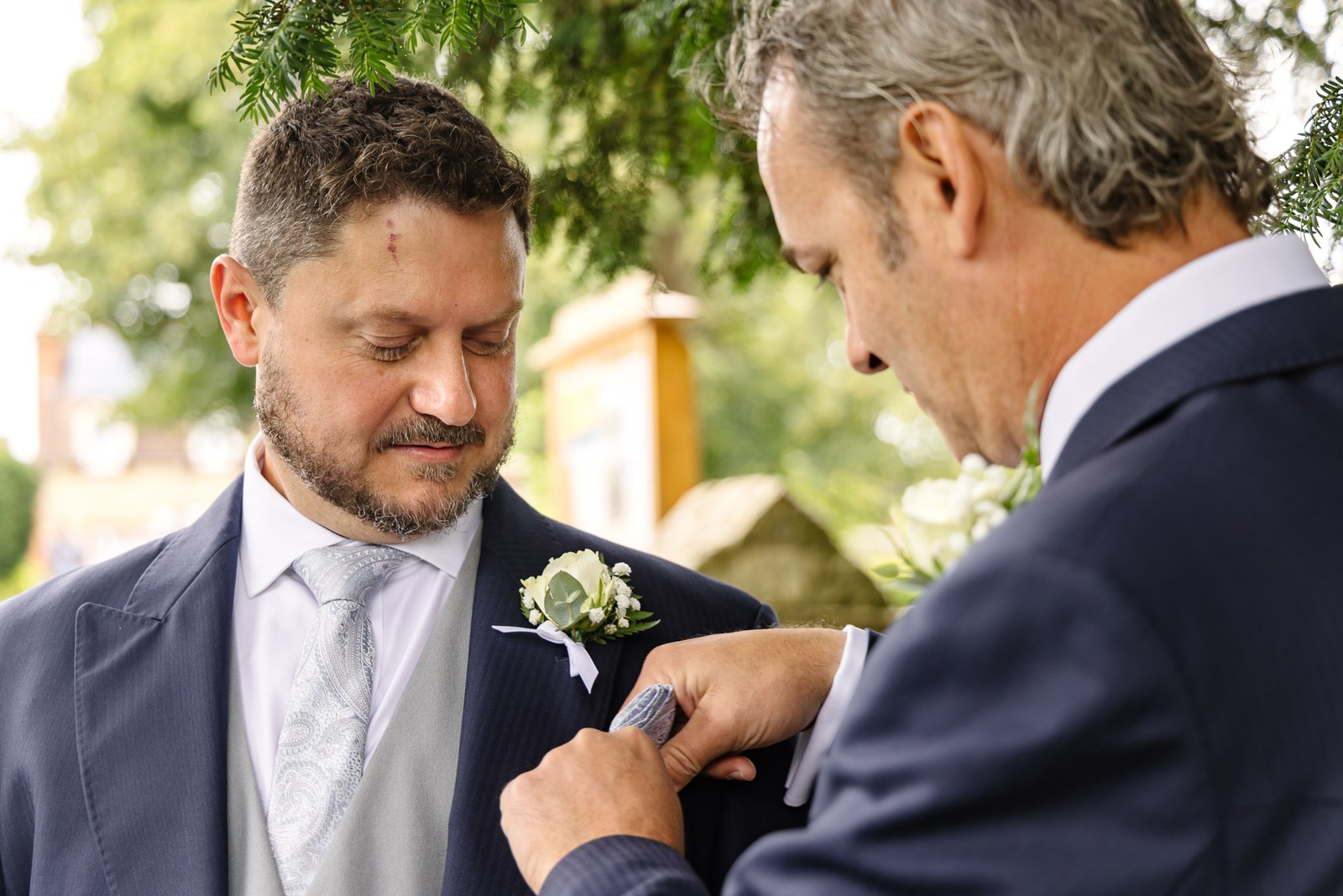 An older man tucks a pocket square into the groom's breast pocket outdoors