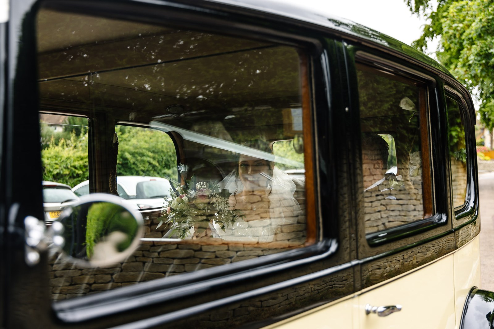 The bride seen through the window of the vintage cream-and-black wedding car, her bouquet beside her