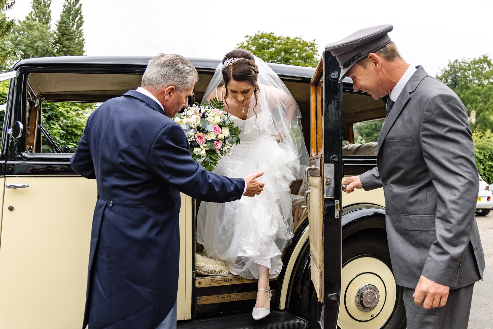The bride is helped from the vintage car by the chauffeur and her father