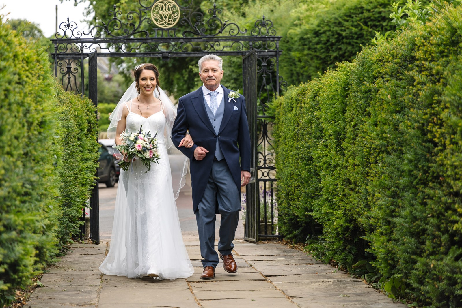 The bride and her father walk through a wrought-iron gate in a tall hedge toward the church