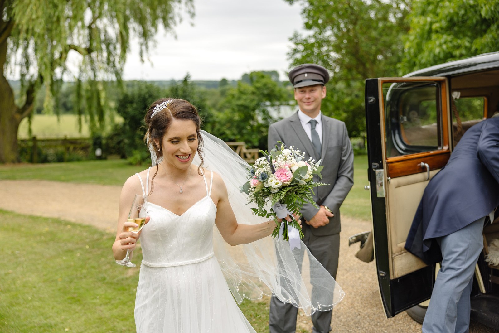 The bride carries a glass of champagne past the vintage car and its chauffeur