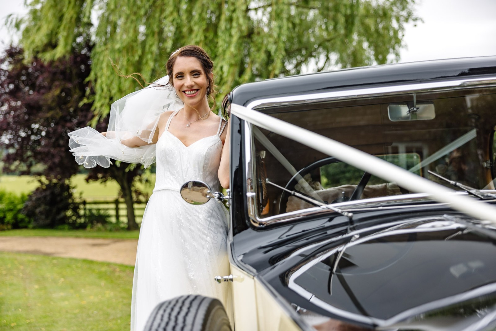 The bride leans against the vintage car, her veil catching the breeze