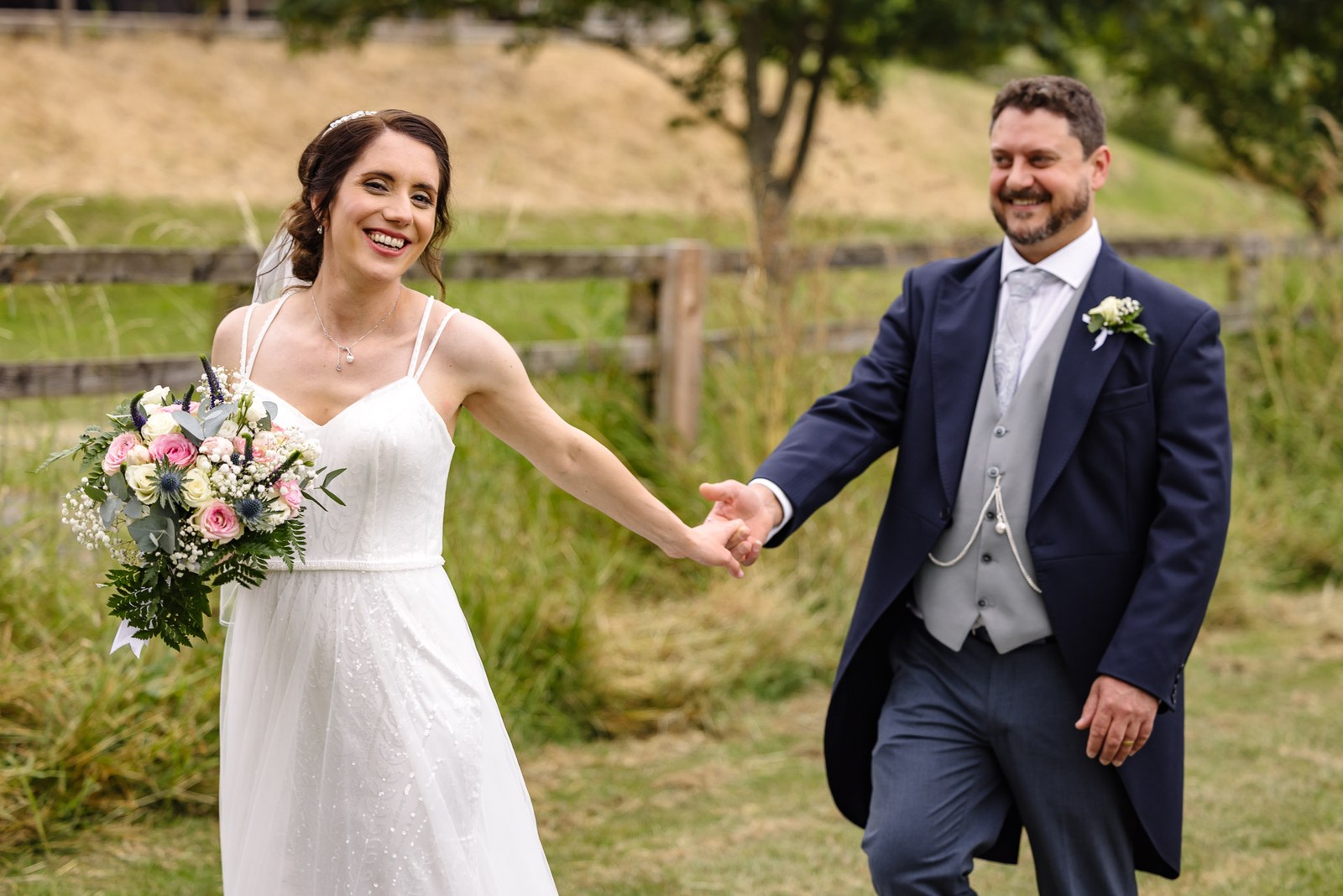 Bride and groom walk hand in hand through a hay meadow by a fence