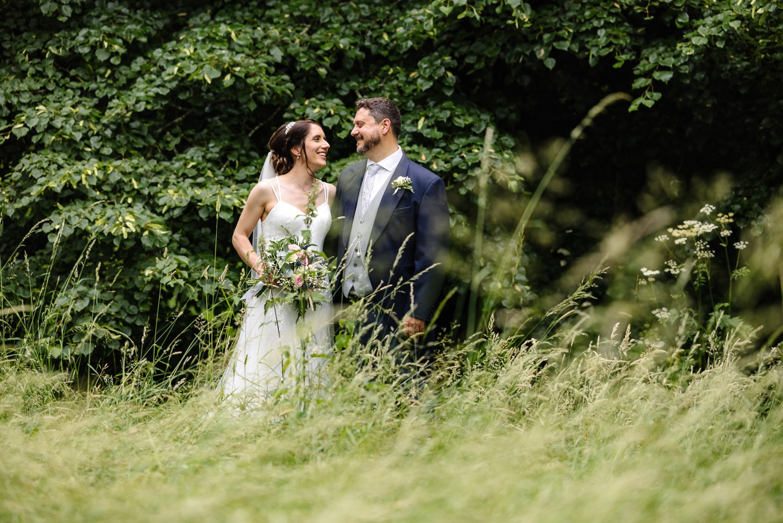 The couple stand close together amid tall grass and greenery