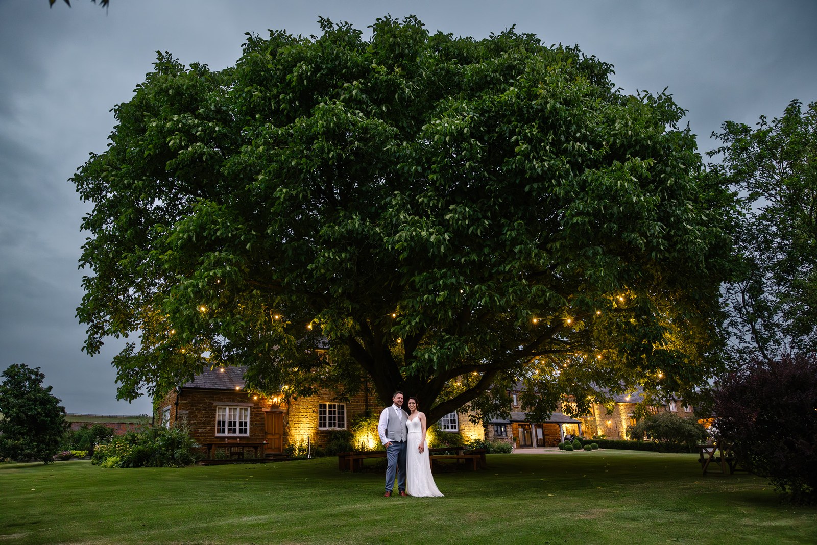 The couple stand beneath a great fairy-lit tree beside the farmhouse at Crockwell Farm at dusk