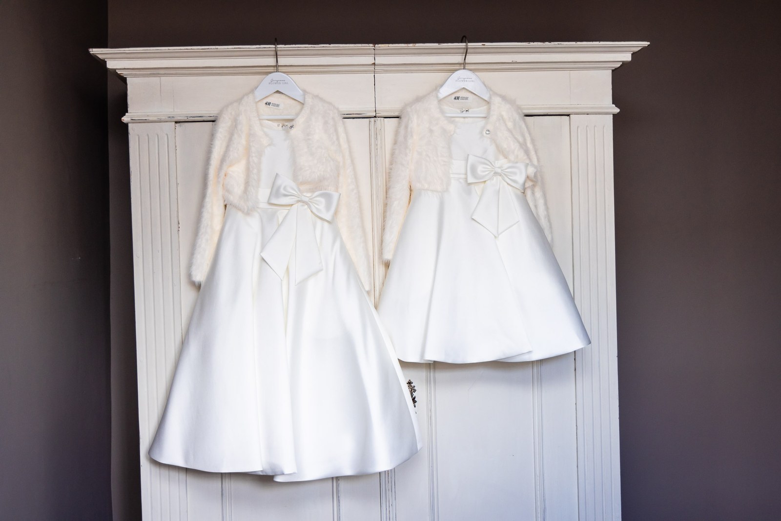 Two flower girl dresses with fur boleros hanging on a painted wardrobe