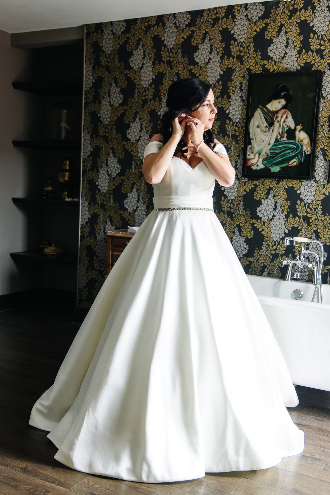 The bride fastens an earring in front of patterned wallpaper in her full gown