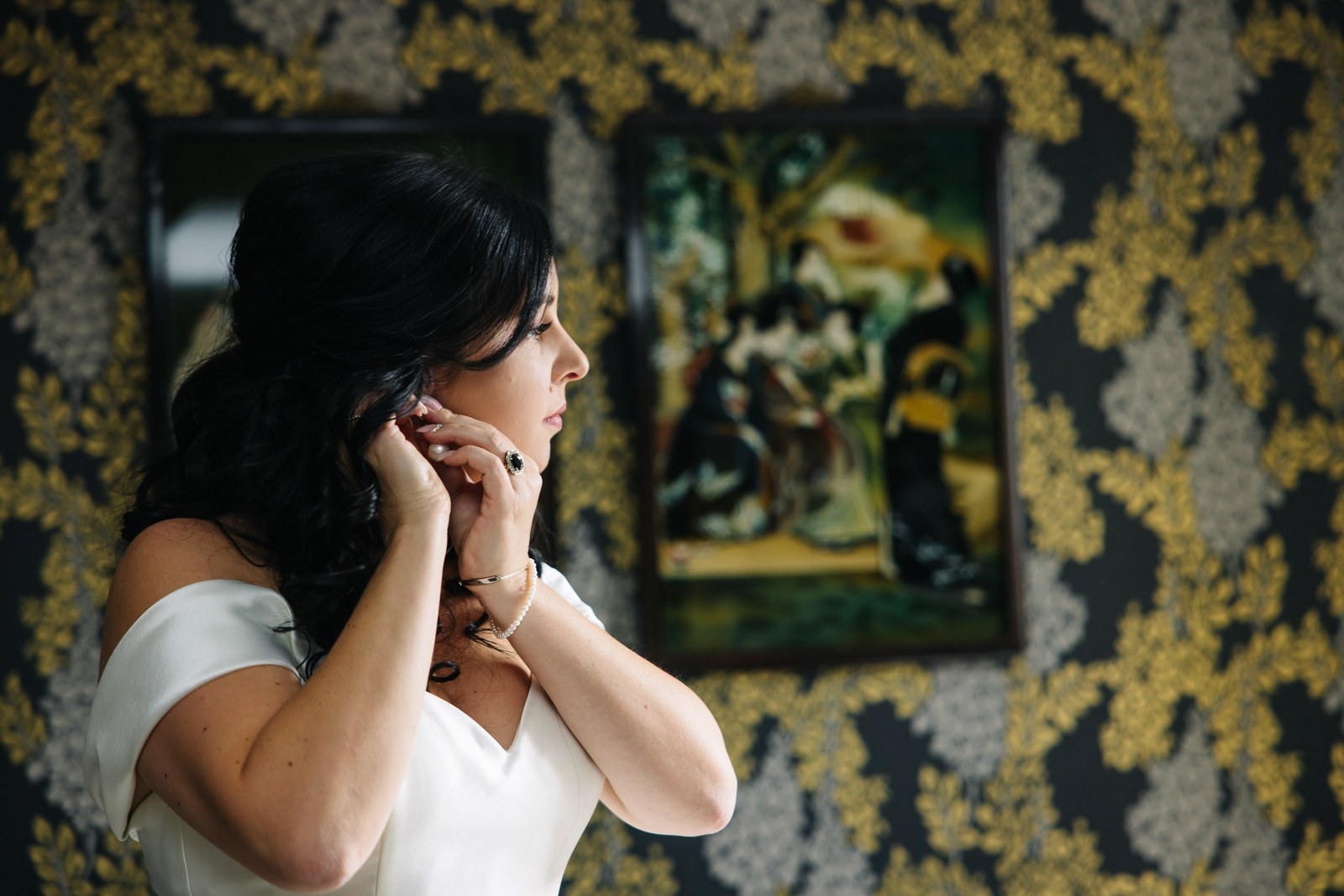 The bride adjusts her earring beside a framed picture on golden wallpaper