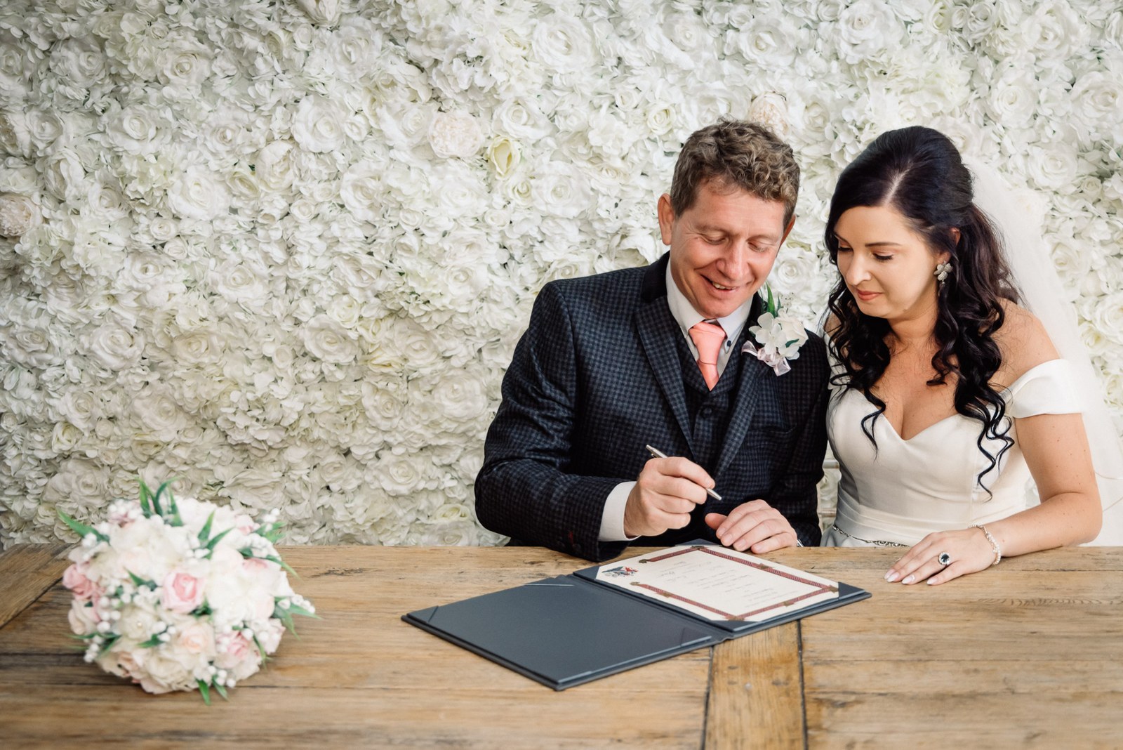 The couple sign the register at a wooden table before the flower wall