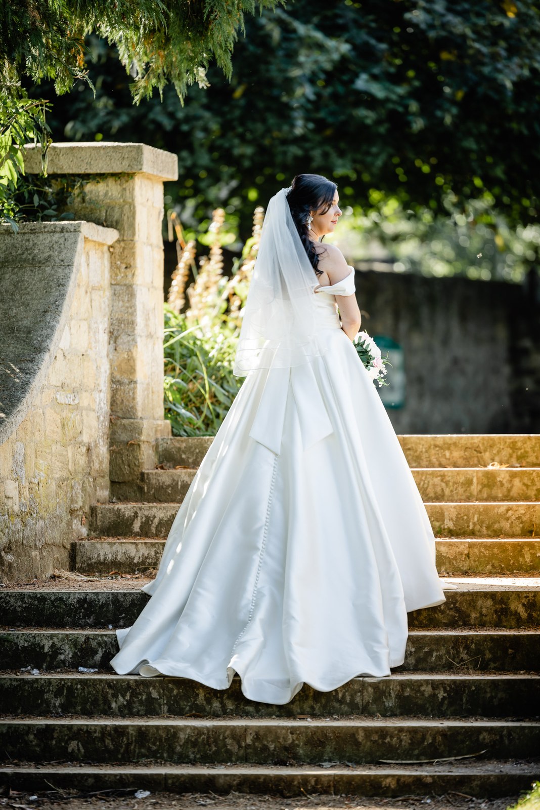 The bride climbs weathered stone steps, her full gown and veil trailing
