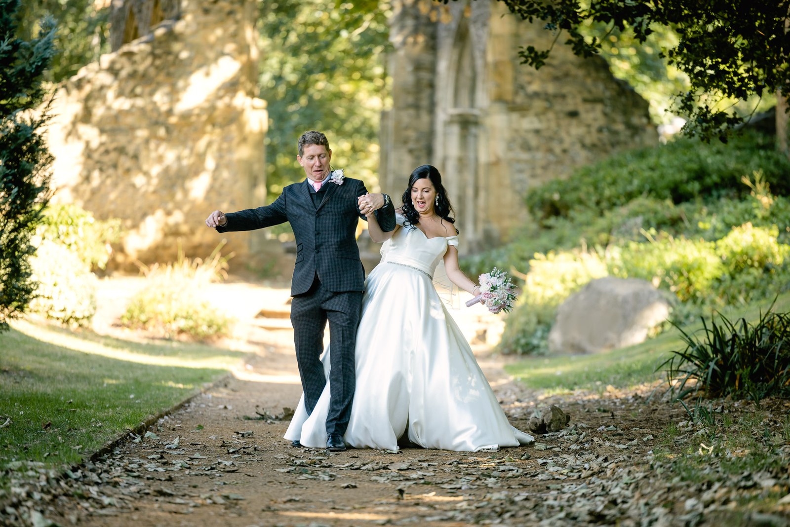 The groom throws his arms wide beside the bride among the abbey ruins