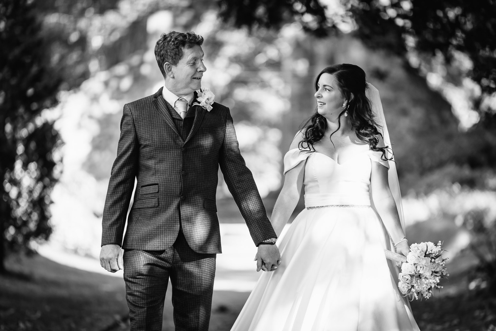 Black-and-white portrait of the couple walking hand in hand beneath trees